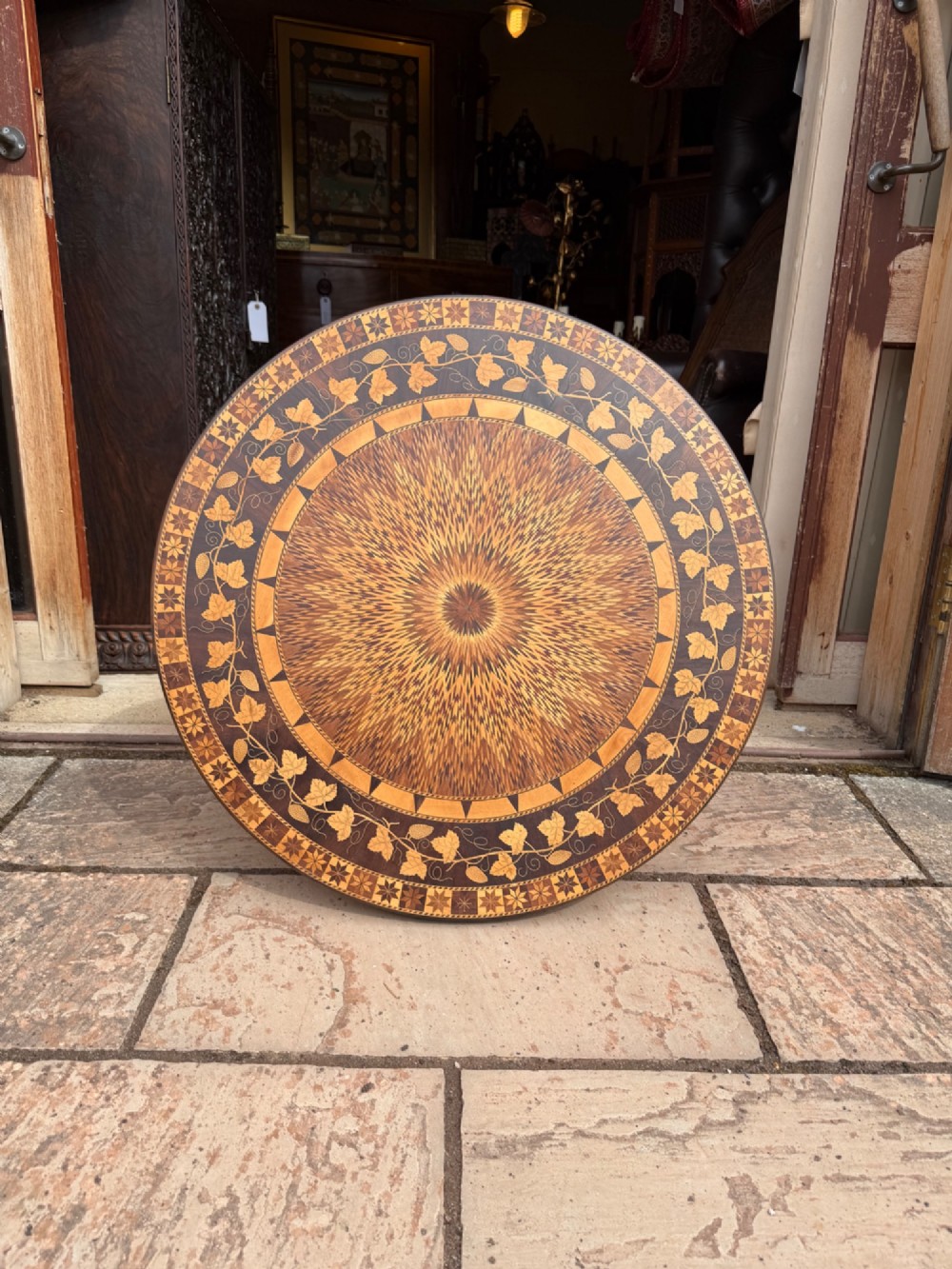 A circular wooden pedestal table featuring elaborate geometric starburst and floral vine marquetry inlays supported by a turned central column and carved tripod legs. View II