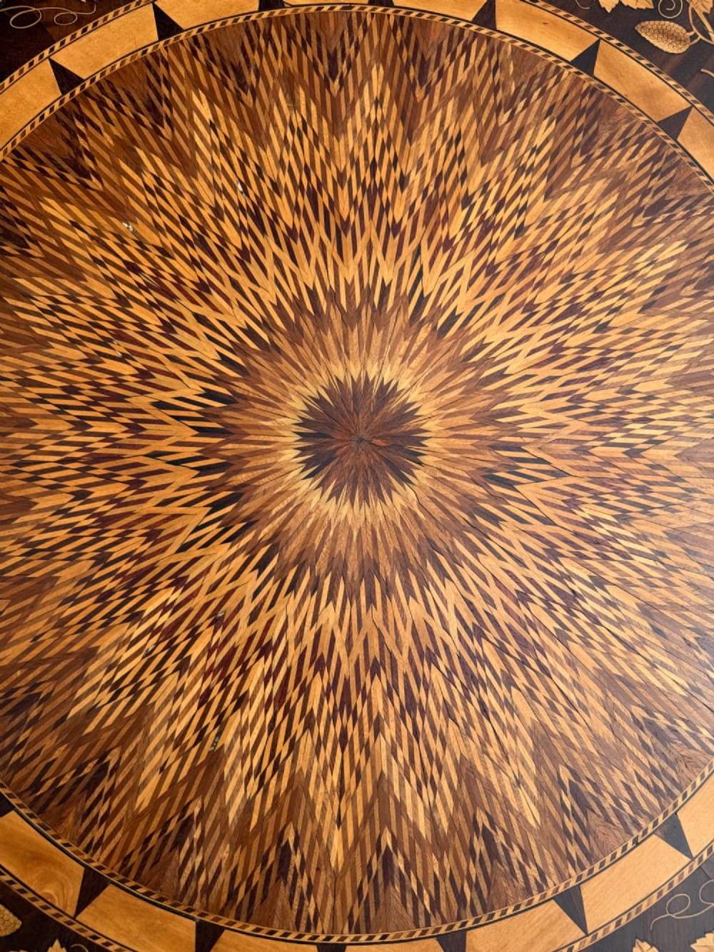 A circular wooden pedestal table featuring elaborate geometric starburst and floral vine marquetry inlays supported by a turned central column and carved tripod legs. View IV