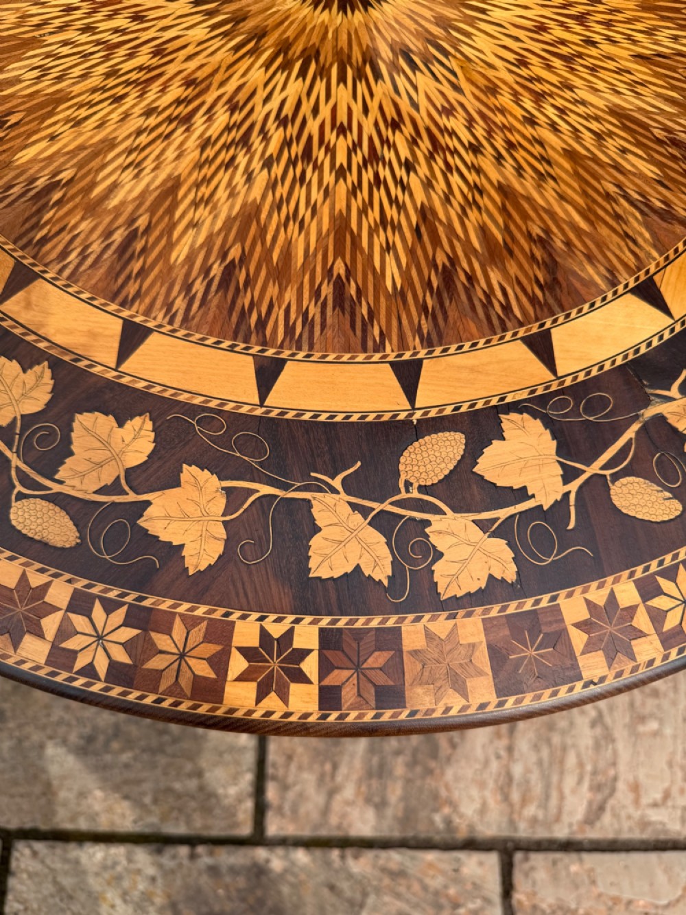 A circular wooden pedestal table featuring elaborate geometric starburst and floral vine marquetry inlays supported by a turned central column and carved tripod legs. View V