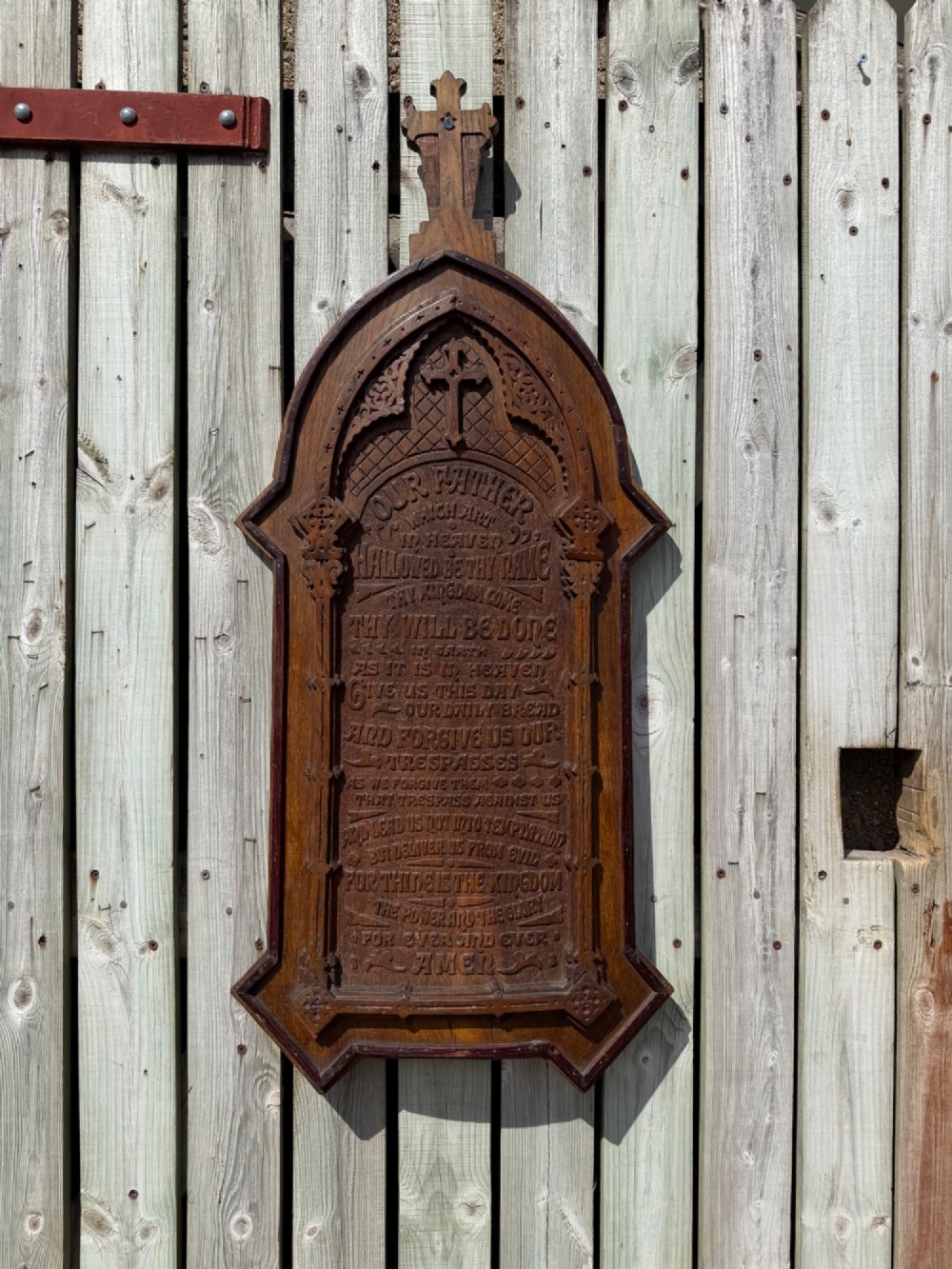 A vertical carved dark oak plaque shaped like a Gothic arch with a cross finial, featuring relief-carved lettering of the Lords Prayer.