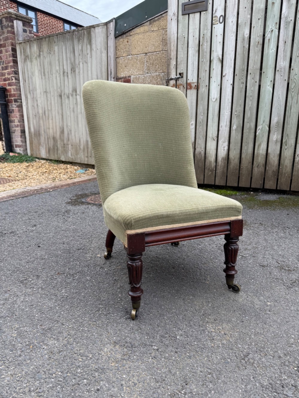 An antique George IV slipper chair upholstered in textured sage green fabric with carved mahogany front legs featuring brass castors.