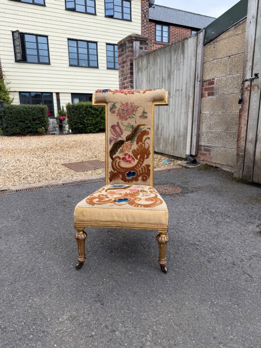 A nineteenth-century prie dieu chair featuring floral needlepoint upholstery, a low seat, a tall T-shaped backrest, and fluted giltwood legs on brass castors.