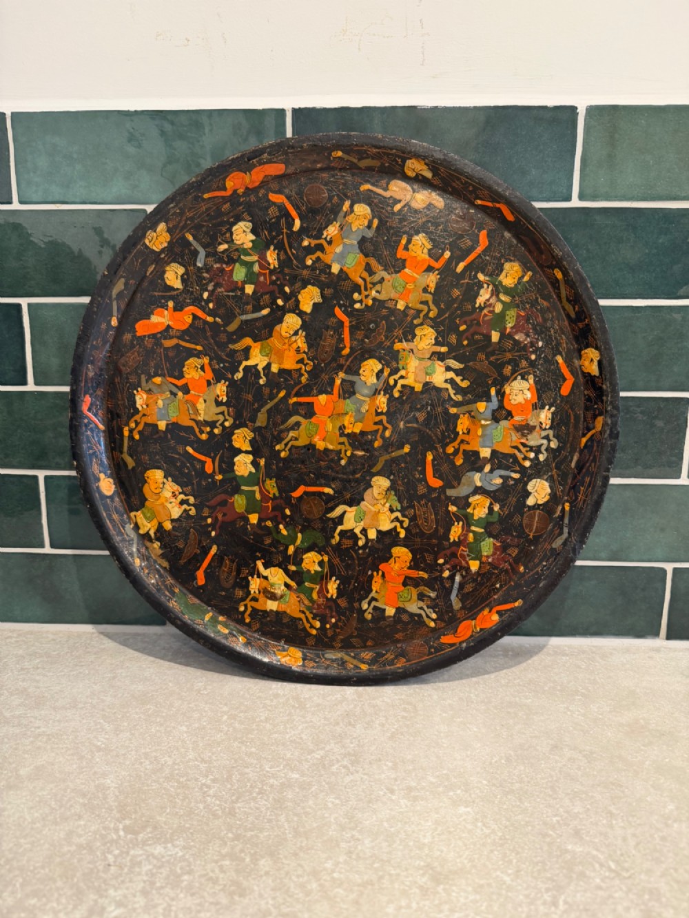 A circular black papier-mache tray hand-painted with equestrian riders in orange, green, and gold against a dark background.