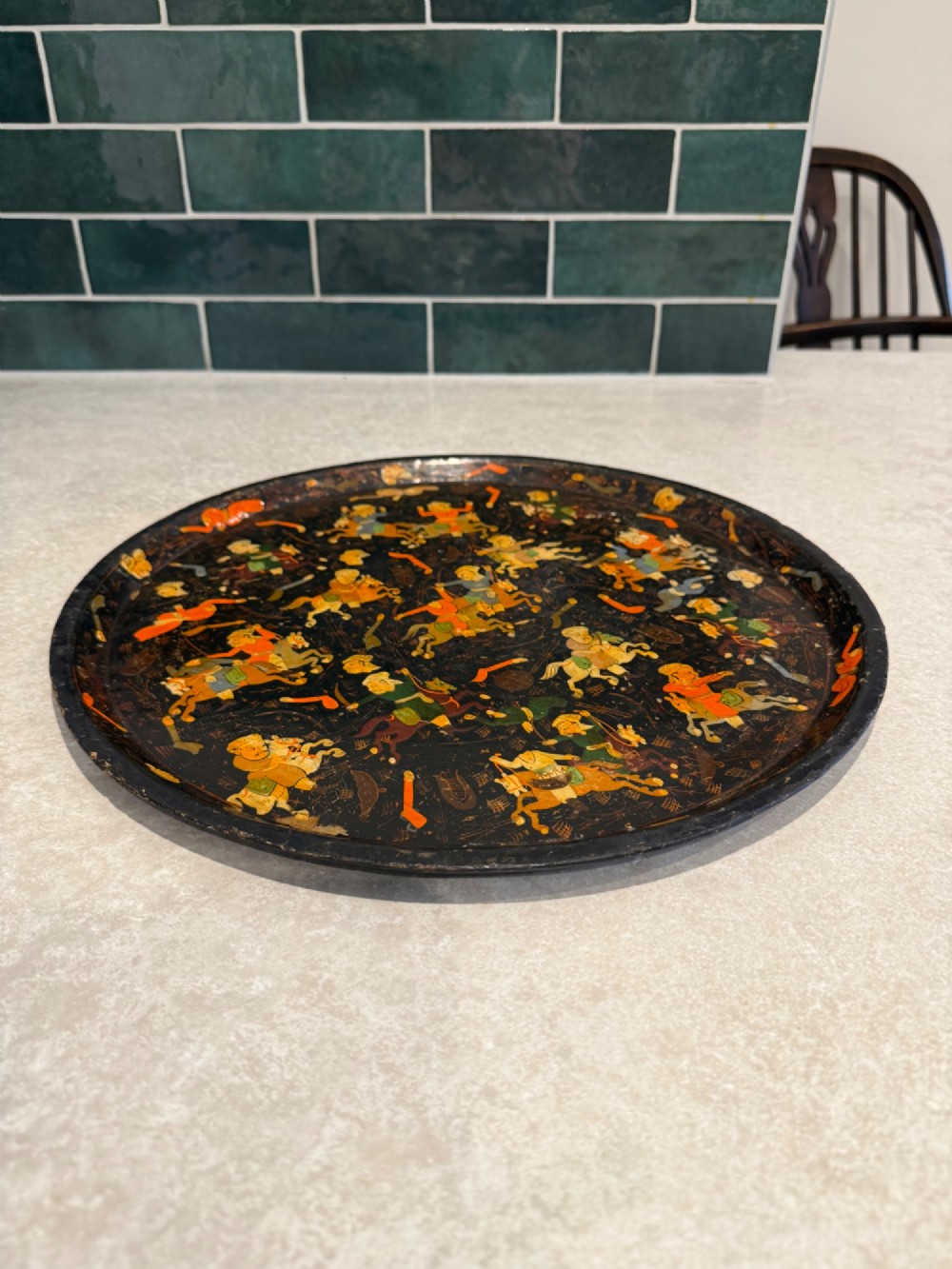 A circular black papier-mache tray hand-painted with equestrian riders in orange, green, and gold against a dark background. View III