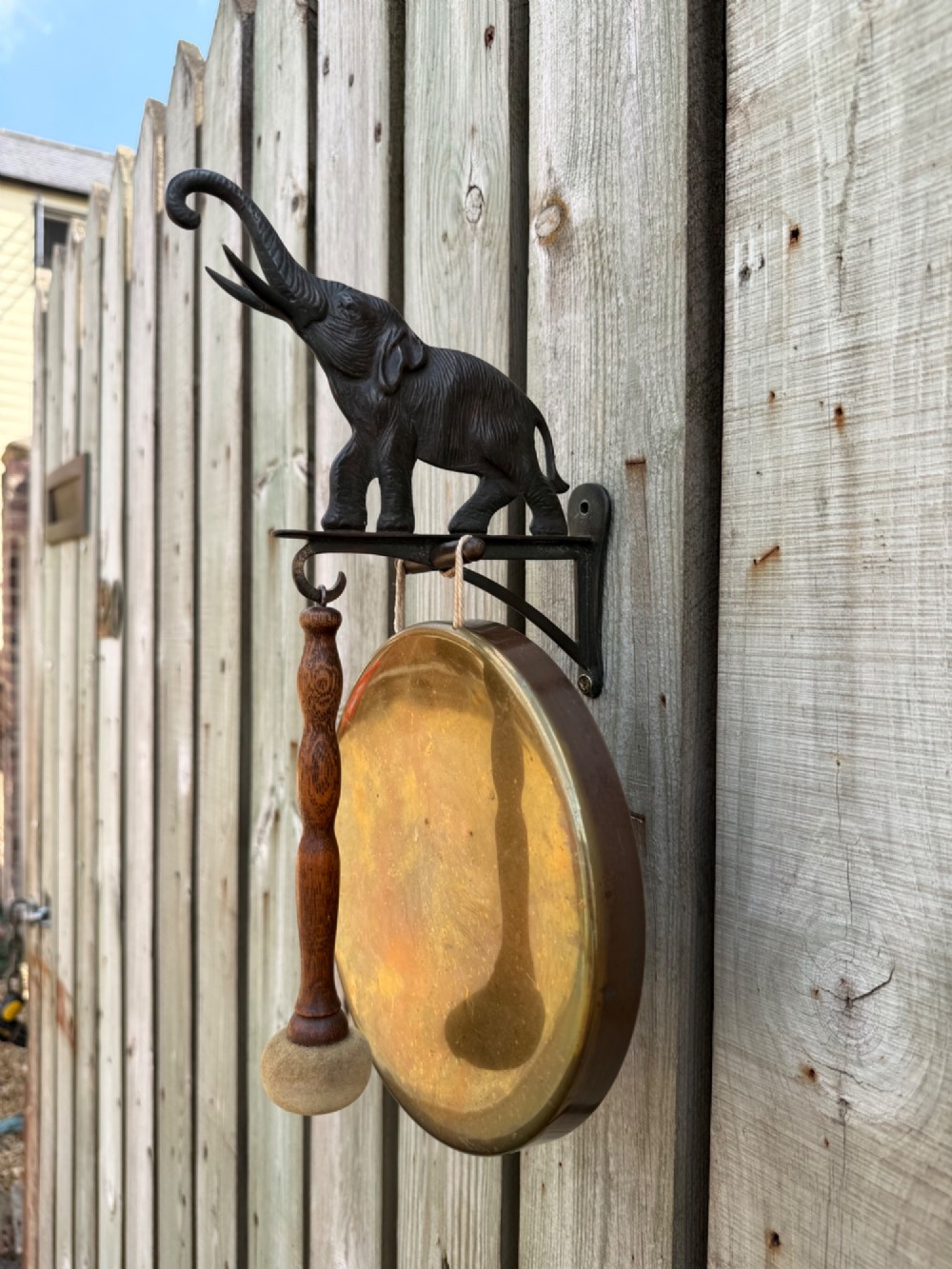 A wall-mounted brass dinner gong suspended from a dark cast iron bracket featuring a detailed elephant figurine, alongside a wooden striker.