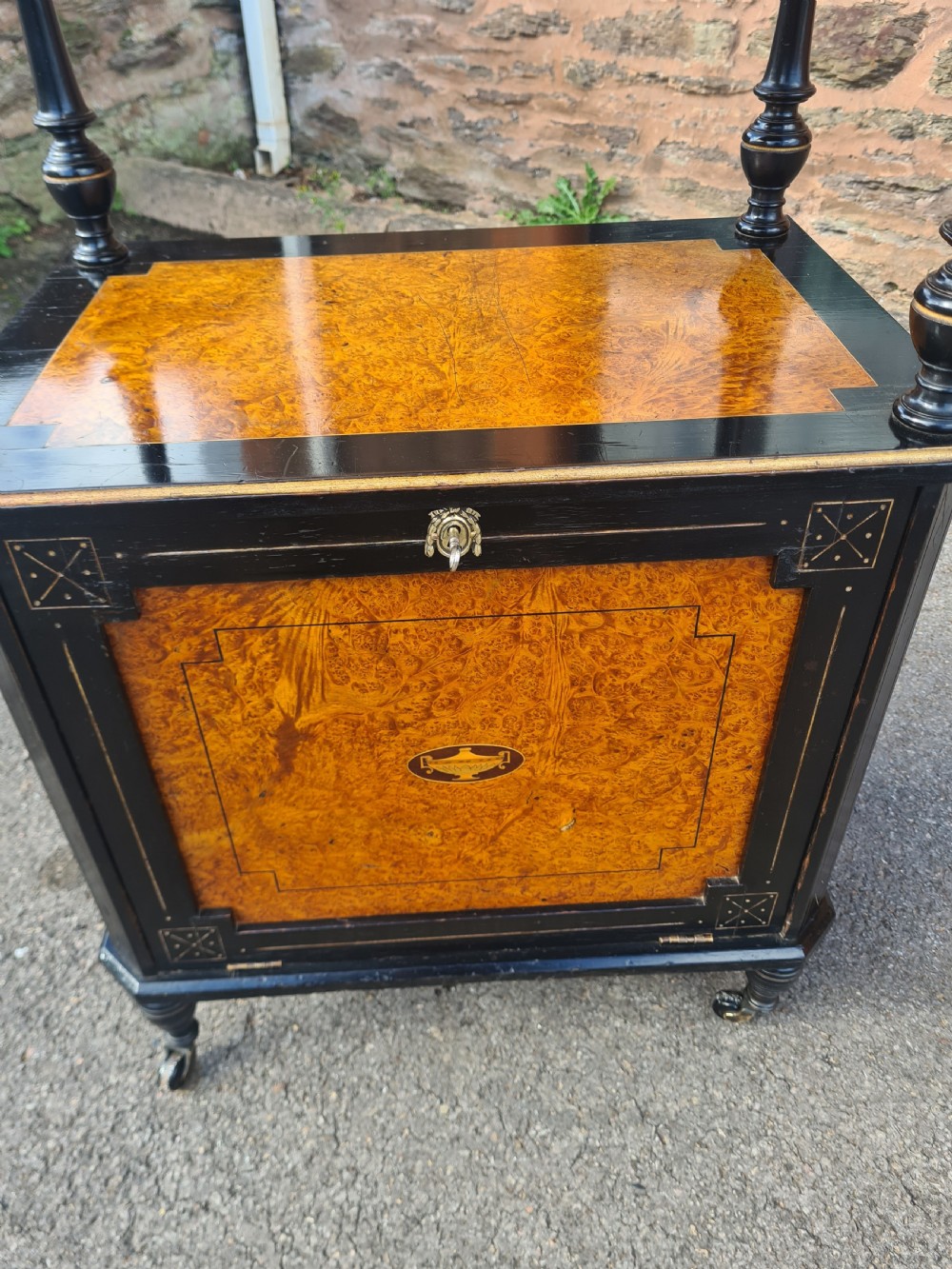 A Victorian ebonised and amboyna wood music cabinet featuring turned columns, two tiers, an inlaid lower cupboard door, and small casters. View V