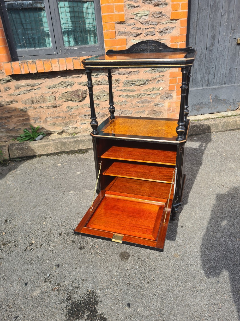 A Victorian ebonised and amboyna wood music cabinet featuring turned columns, two tiers, an inlaid lower cupboard door, and small casters. View VI