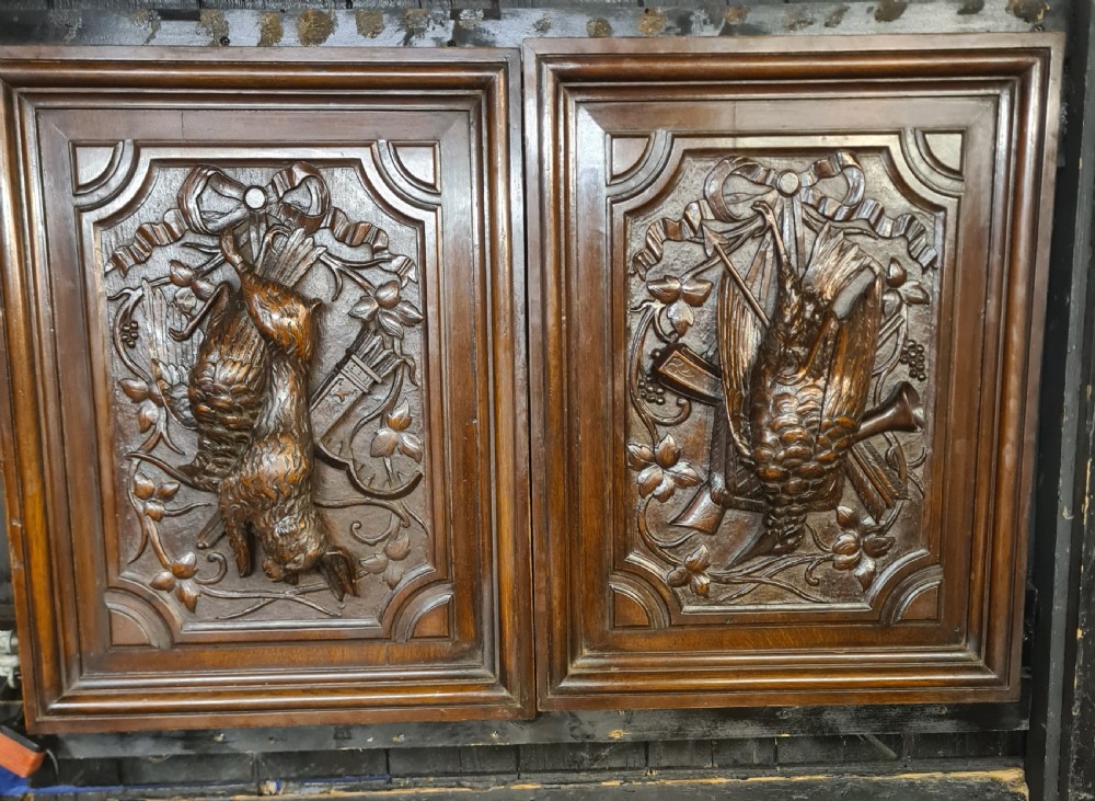 A pair of dark oak timber panels featuring high-relief relief carvings of game birds, a rabbit, ribbons, and hunting equipment.