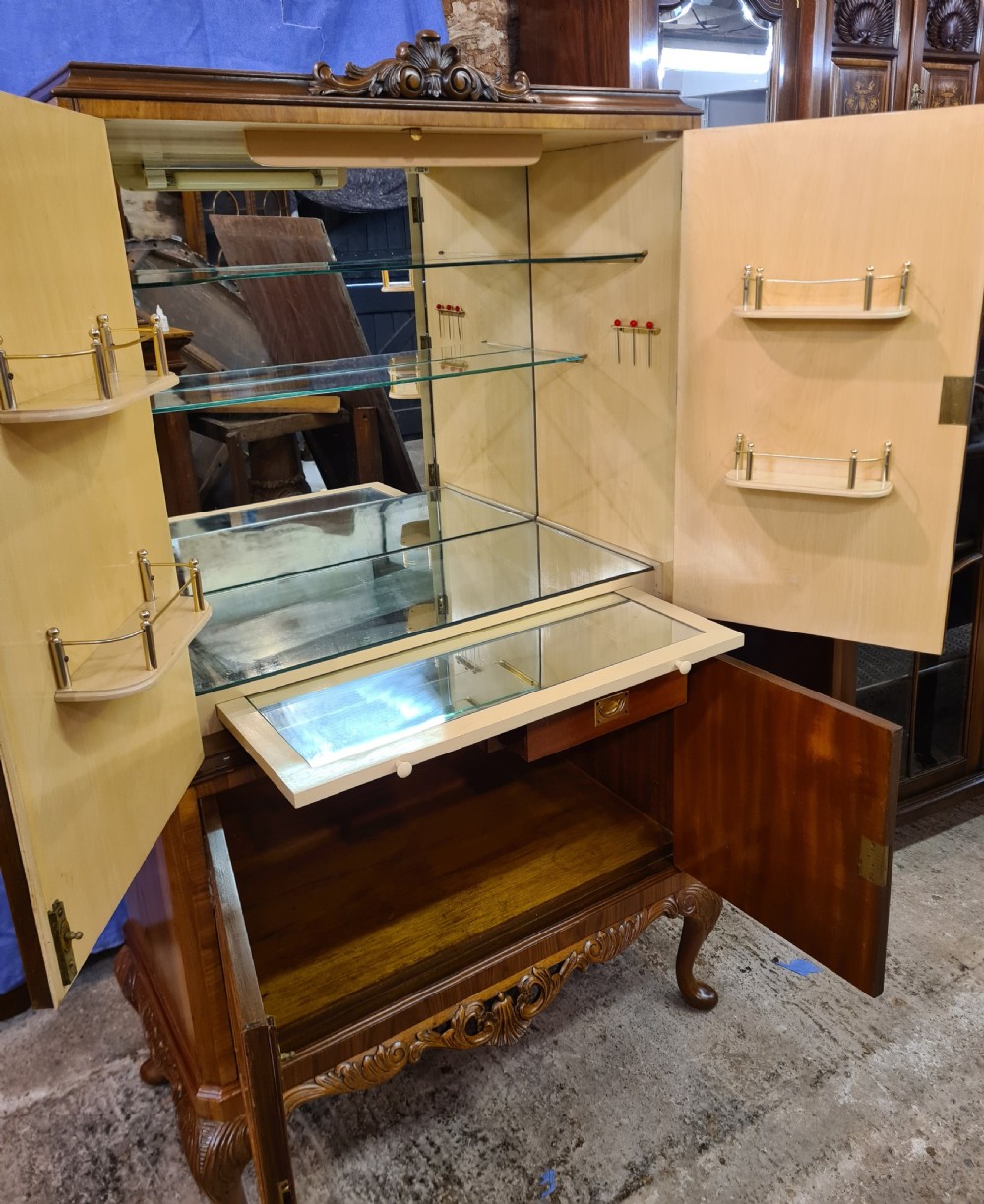 A two-door 1920s burr walnut cocktail cabinet featuring decorative brass strap hinges, book-matched wood grain, a carved apron, and cabriole legs. View VII