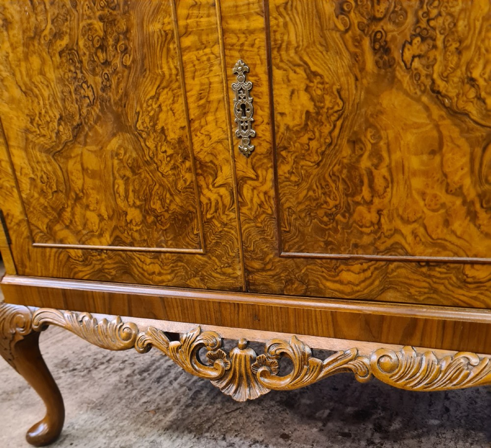 A two-door 1920s burr walnut cocktail cabinet featuring decorative brass strap hinges, book-matched wood grain, a carved apron, and cabriole legs. View XII