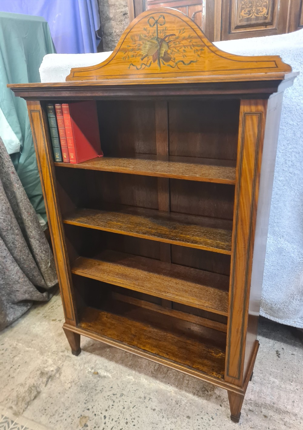 A Victorian satinwood open bookcase featuring four tiered shelves and an arched pediment decorated with marquetry foliage and musical instruments. View VI
