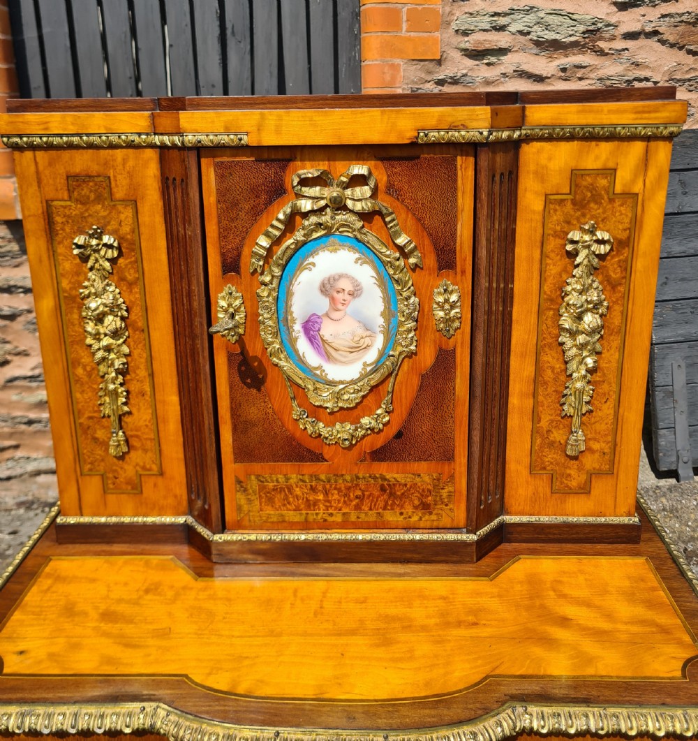 A French satinwood and amboyna bonheur de jour desk featuring ornate gilt-bronze mounts, an inlaid Greek key drawer pattern, and a central cabinet door set with a hand-painted porcelain portrait plaque. View X