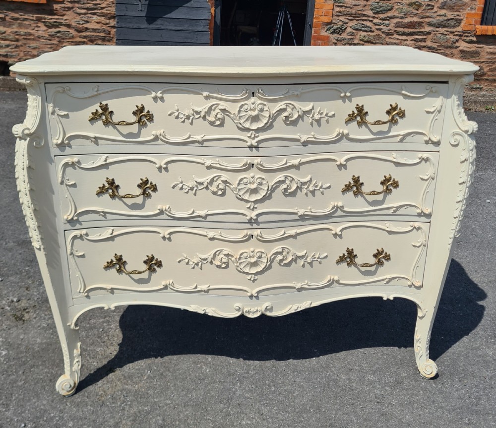 A nineteenth-century cream painted serpentine commode featuring three drawers with rococo style relief carvings and brass handles.