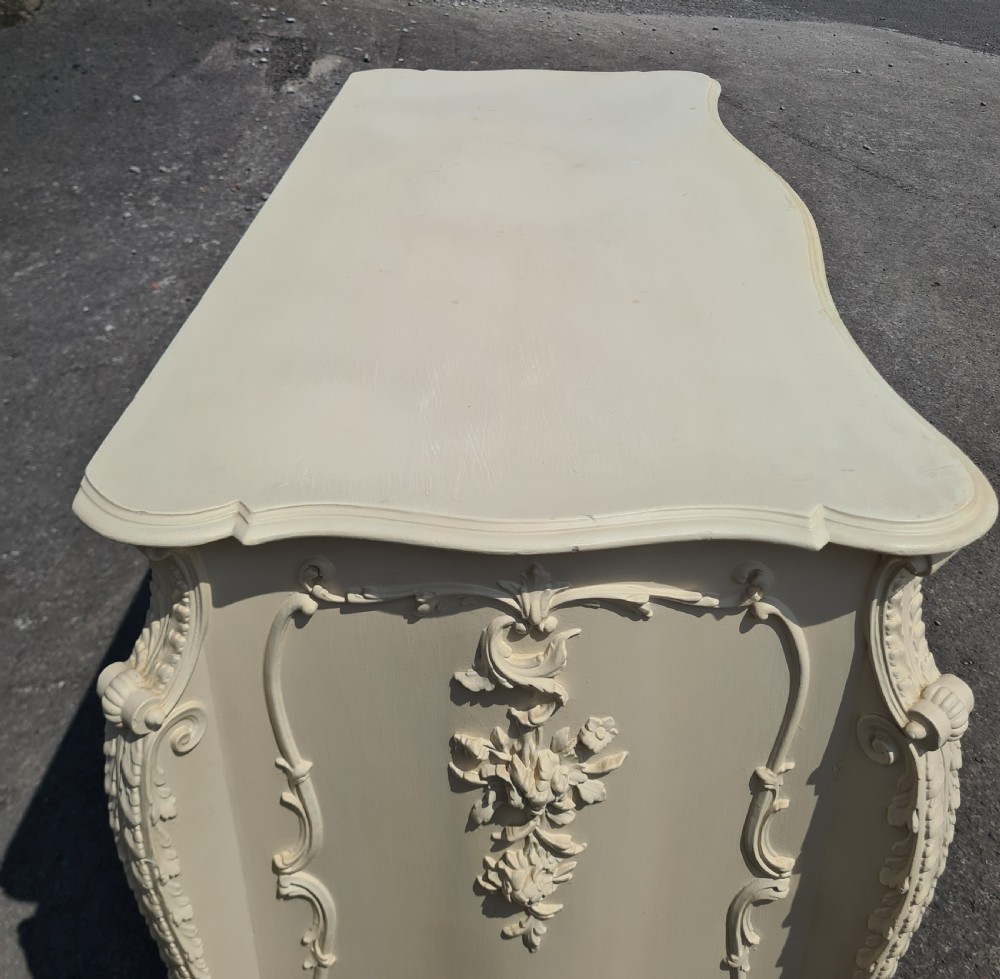 A nineteenth-century cream painted serpentine commode featuring three drawers with rococo style relief carvings and brass handles. View VI