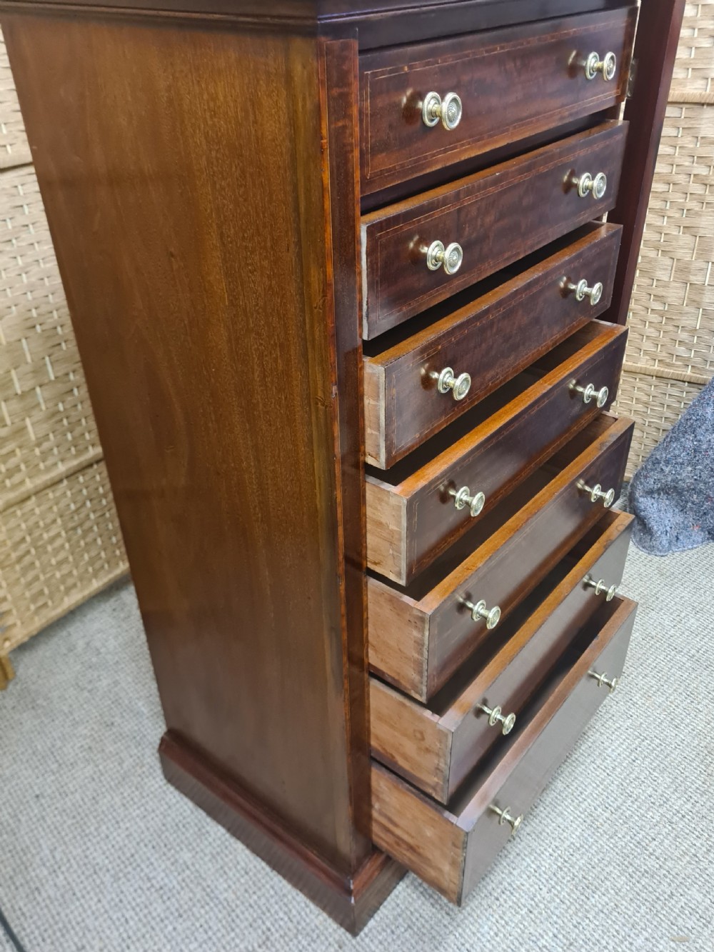 A tall, narrow late Victorian mahogany Wellington chest featuring seven drawers with satinwood stringing inlays, round brass knobs, and a side locking bar. View VI