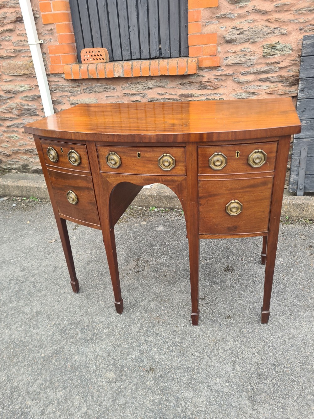 A Georgian mahogany bowfront sideboard featuring a curved front, octagonal brass handles, three drawers, and four square tapered legs. View IV