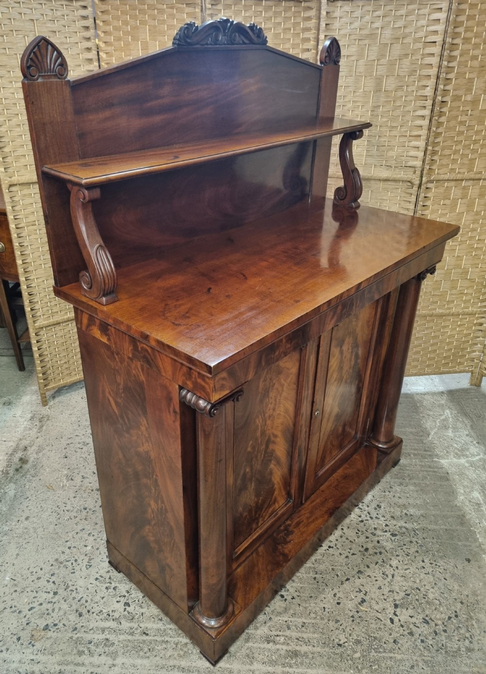 An early nineteenth-century flame mahogany chiffonier featuring a carved cresting backboard with a single shelf over a two-door cabinet flanked by turned columns. View III