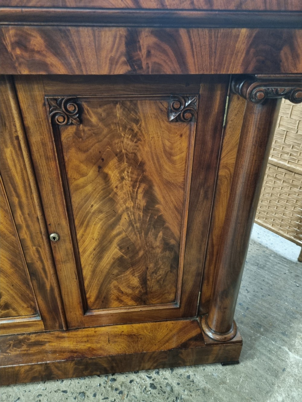 An early nineteenth-century flame mahogany chiffonier featuring a carved cresting backboard with a single shelf over a two-door cabinet flanked by turned columns. View IV