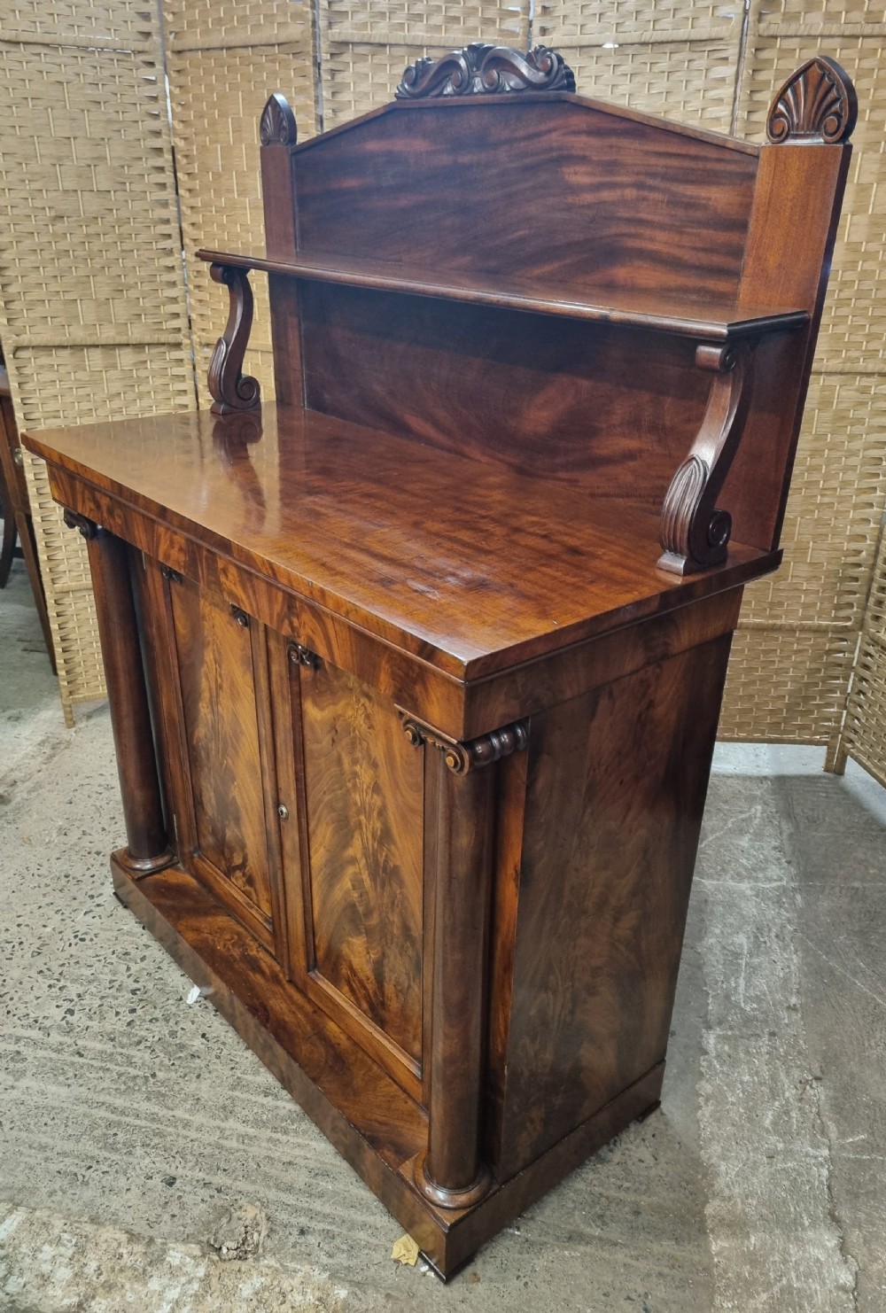 An early nineteenth-century flame mahogany chiffonier featuring a carved cresting backboard with a single shelf over a two-door cabinet flanked by turned columns. View VIII