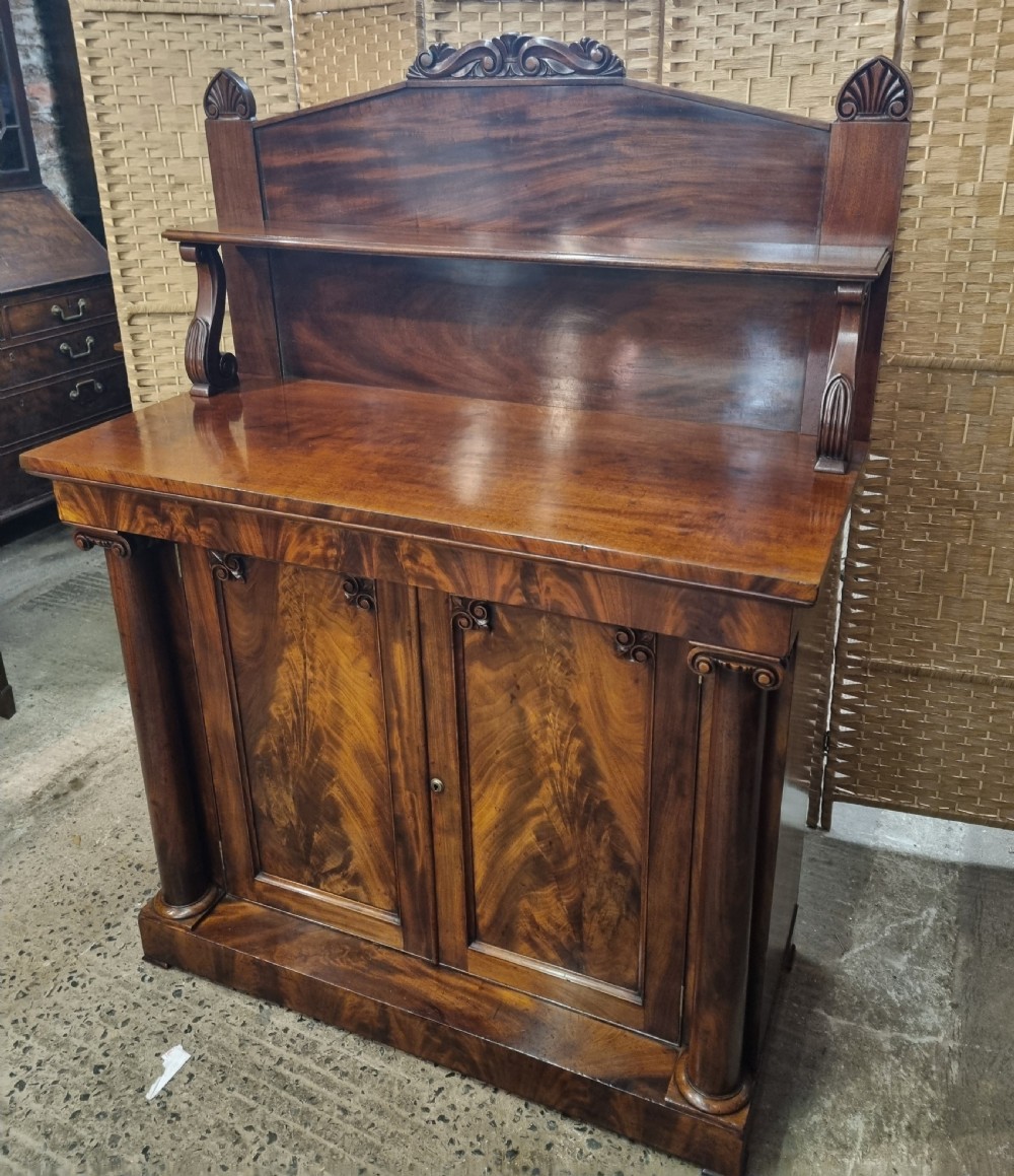 An early nineteenth-century flame mahogany chiffonier featuring a carved cresting backboard with a single shelf over a two-door cabinet flanked by turned columns. View X