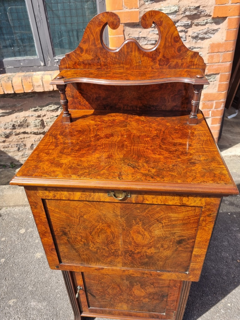 An Edwardian burr walnut music cabinet with a decorative scrolled upstand, a top shelf supported by turned pillars, a fall-front main compartment, and a lower single door cupboard. View II