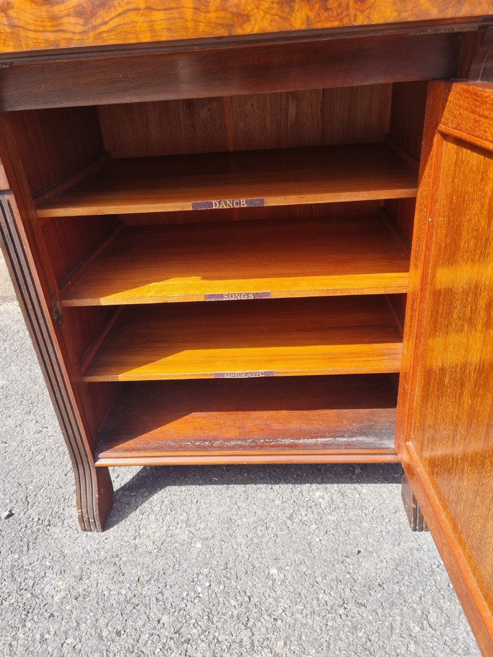 An Edwardian burr walnut music cabinet with a decorative scrolled upstand, a top shelf supported by turned pillars, a fall-front main compartment, and a lower single door cupboard. View VI