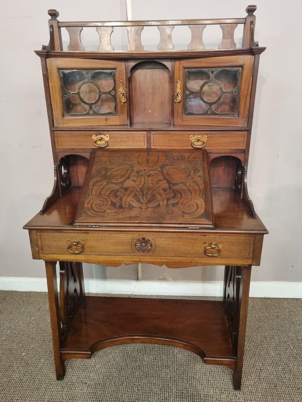 An Arts and Crafts mahogany bureau cabinet featuring a carved fall front slope, glazed upper cupboard doors, brass handles, and a lower shelf.