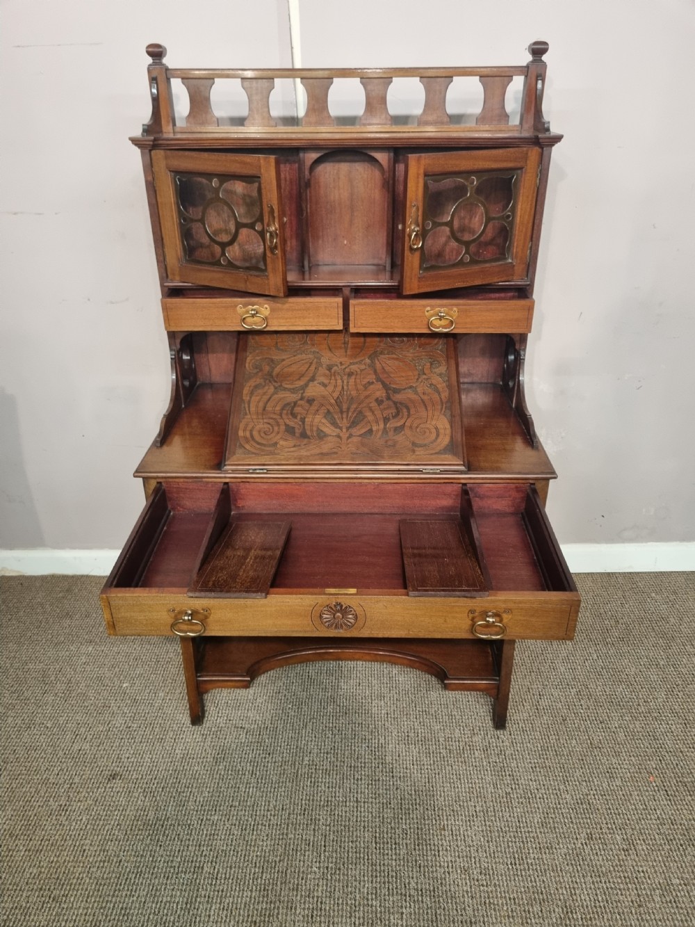 An Arts and Crafts mahogany bureau cabinet featuring a carved fall front slope, glazed upper cupboard doors, brass handles, and a lower shelf. View III
