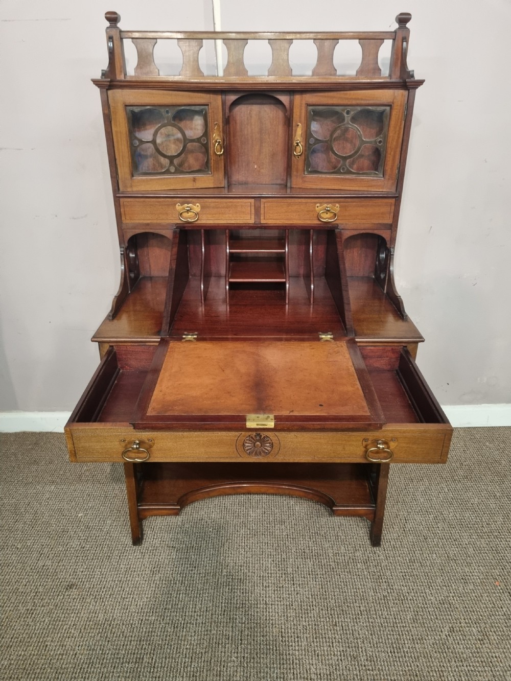 An Arts and Crafts mahogany bureau cabinet featuring a carved fall front slope, glazed upper cupboard doors, brass handles, and a lower shelf. View IV