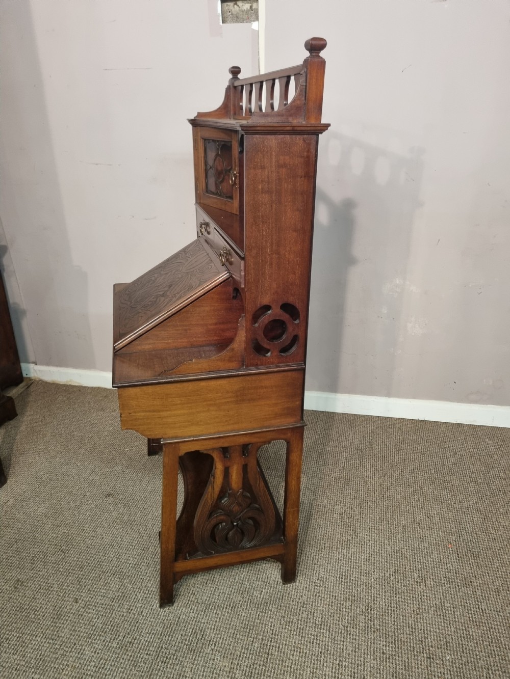 An Arts and Crafts mahogany bureau cabinet featuring a carved fall front slope, glazed upper cupboard doors, brass handles, and a lower shelf. View V