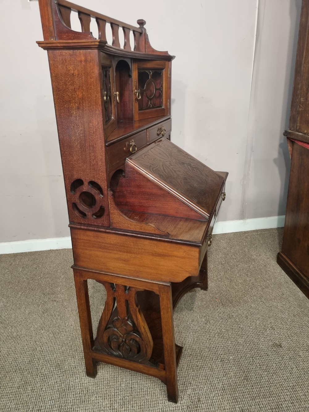 An Arts and Crafts mahogany bureau cabinet featuring a carved fall front slope, glazed upper cupboard doors, brass handles, and a lower shelf. View VII