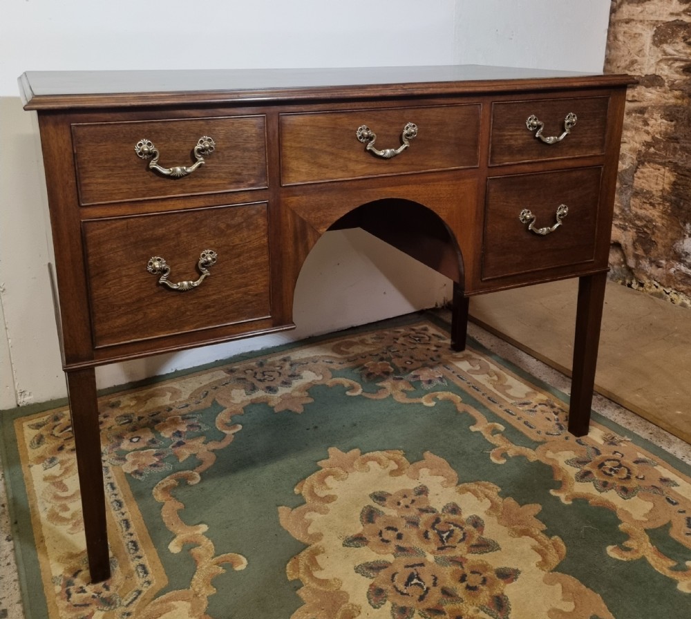 A Georgian Revival mahogany kneehole dressing table featuring five drawers with ornate brass drop handles and an arched central apron.