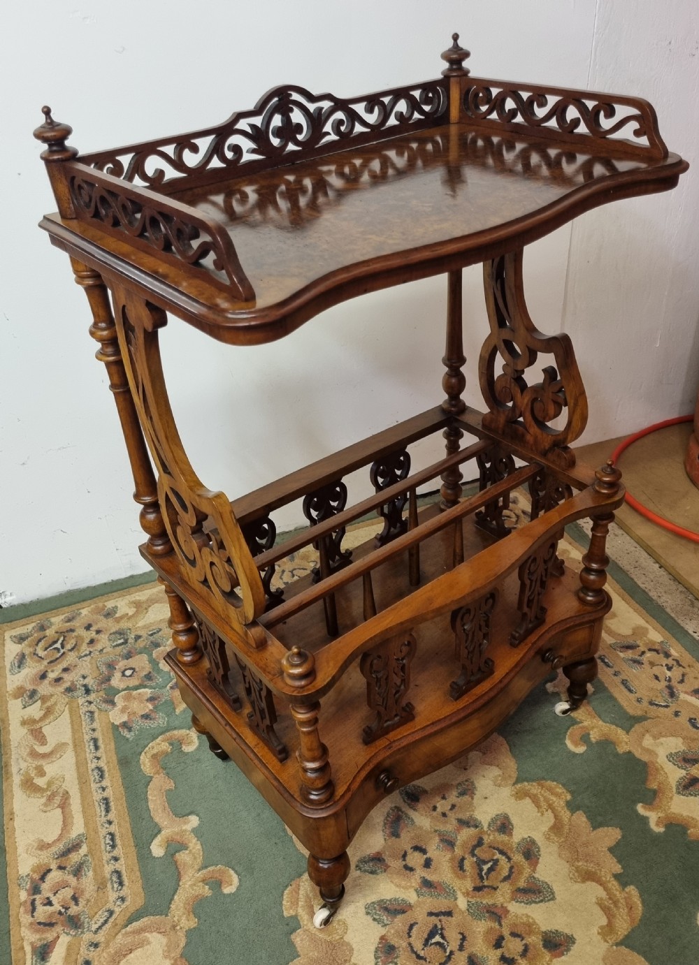 A Victorian burr walnut Canterbury featuring a serpentine top shelf with a carved pierced gallery, vertical divided compartments, a lower drawer, and turned legs on castors.