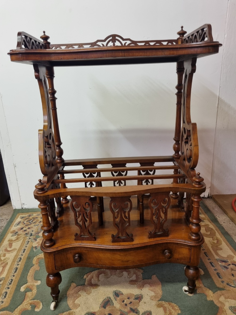 A Victorian burr walnut Canterbury featuring a serpentine top shelf with a carved pierced gallery, vertical divided compartments, a lower drawer, and turned legs on castors. View II