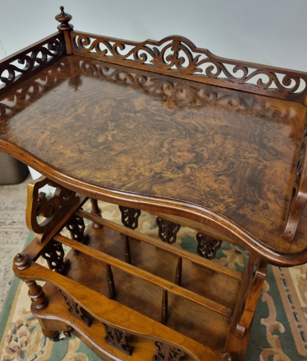 A Victorian burr walnut Canterbury featuring a serpentine top shelf with a carved pierced gallery, vertical divided compartments, a lower drawer, and turned legs on castors. View III