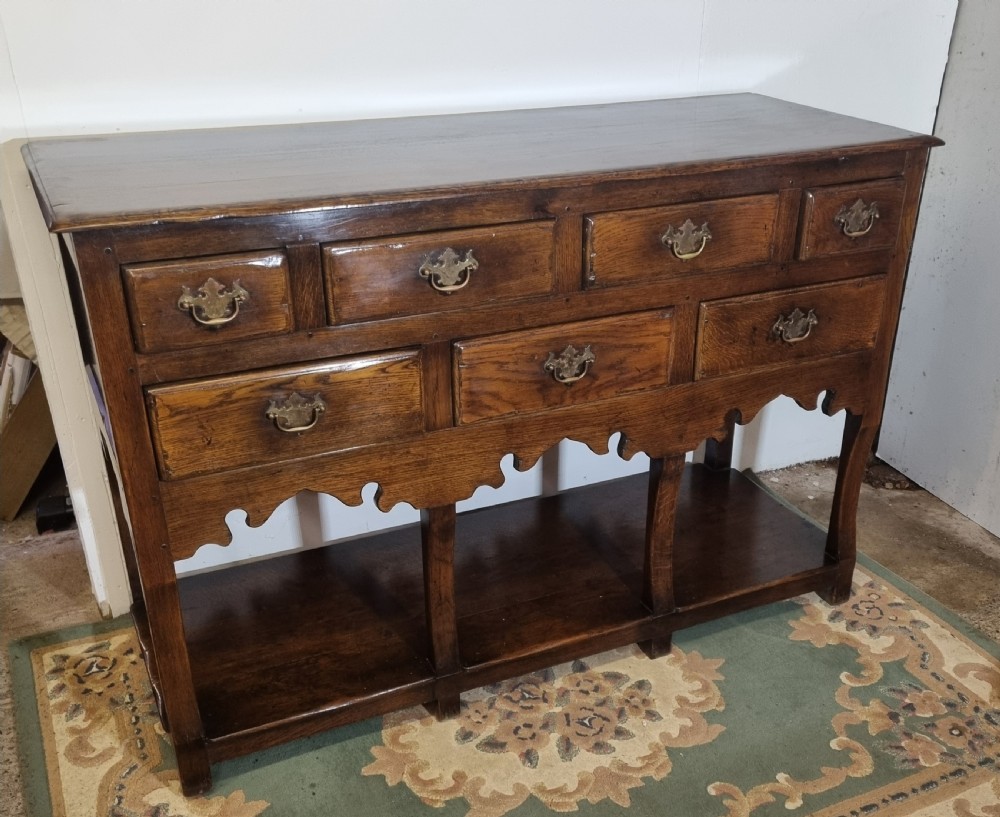 A dark oak Georgian-style dresser base featuring multiple drawers with brass handles, a scalloped apron, and an open lower potboard shelf.