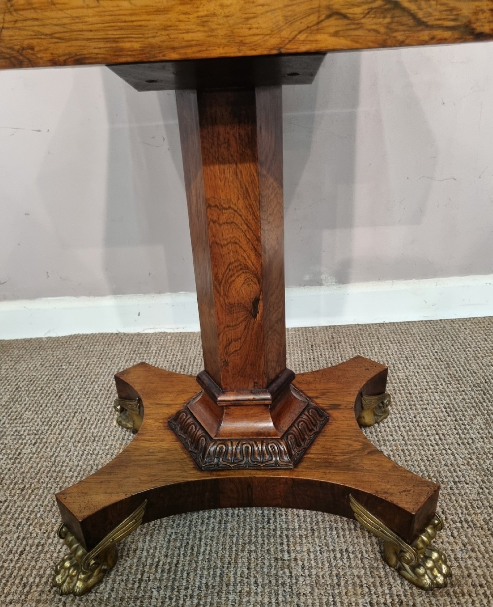 A Regency rosewood teapoy featuring a hinged sarcophagus chest supported by a tapered column and quadform base with brass lion paw feet. View V