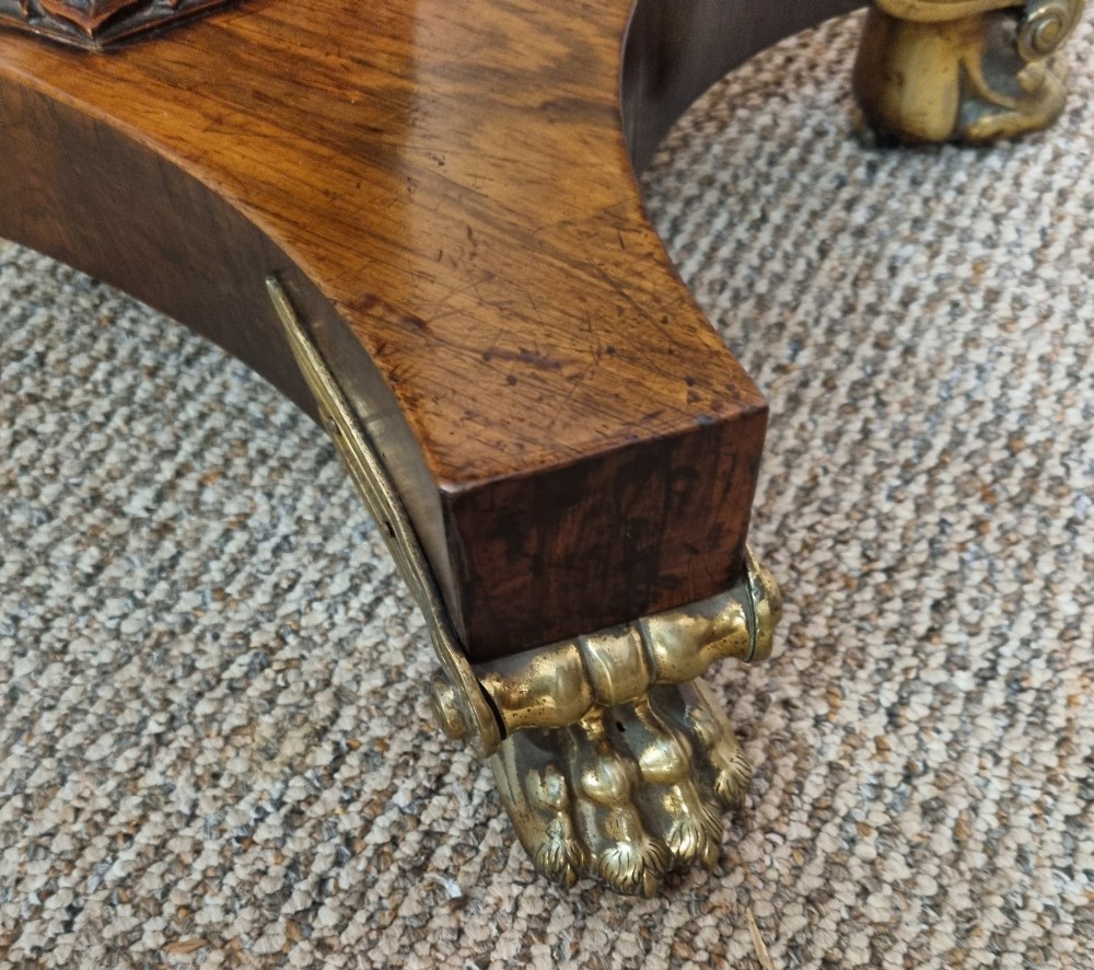 A Regency rosewood teapoy featuring a hinged sarcophagus chest supported by a tapered column and quadform base with brass lion paw feet. View VIII