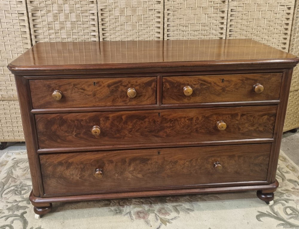 A nineteenth-century flame mahogany chest of drawers featuring two short over two long drawers with turned wooden knobs, rounded moulding, and short turned feet with castors.