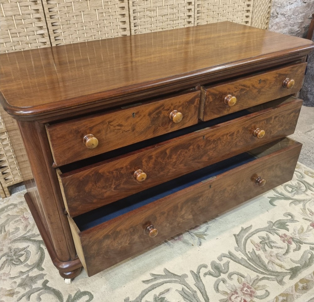 A nineteenth-century flame mahogany chest of drawers featuring two short over two long drawers with turned wooden knobs, rounded moulding, and short turned feet with castors. View II