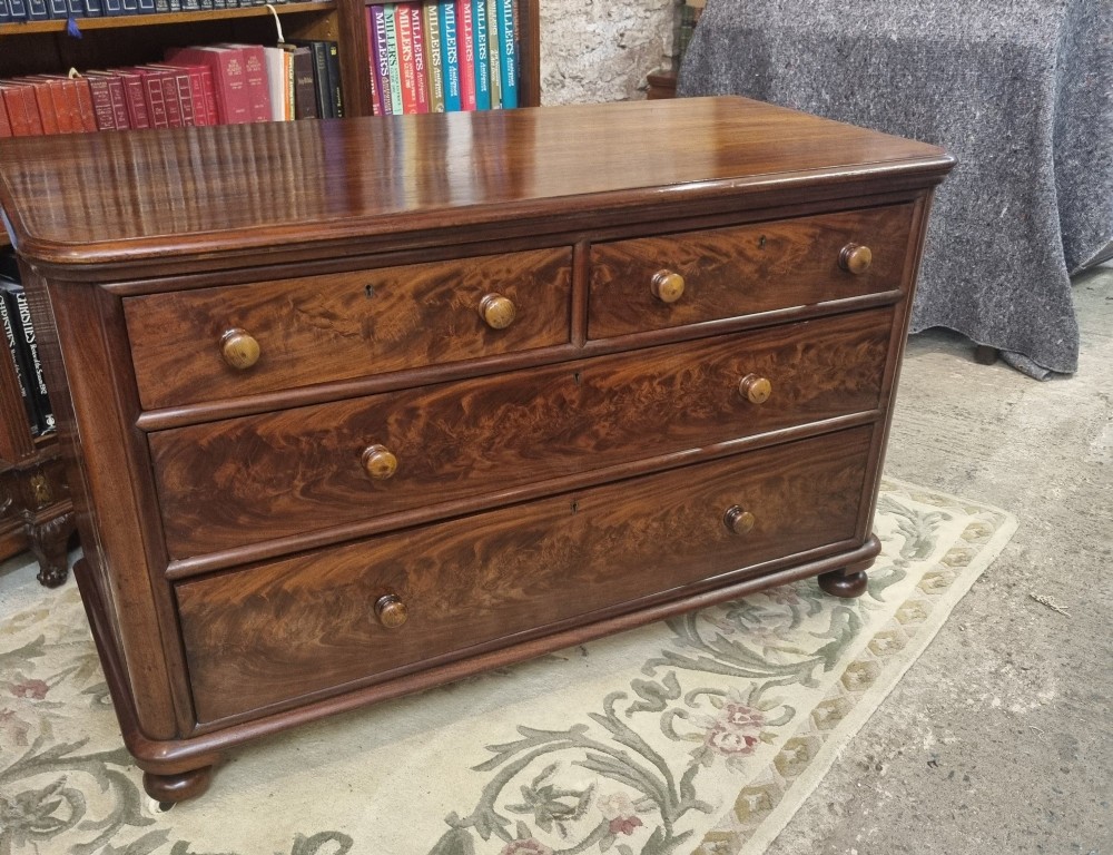A nineteenth-century flame mahogany chest of drawers featuring two short over two long drawers with turned wooden knobs, rounded moulding, and short turned feet with castors. View VIII