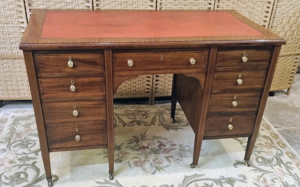 An Edwardian mahogany writing desk featuring nine inlaid drawers, a terracotta leather writing surface, brass knob handles, and tapered legs on castors. View II