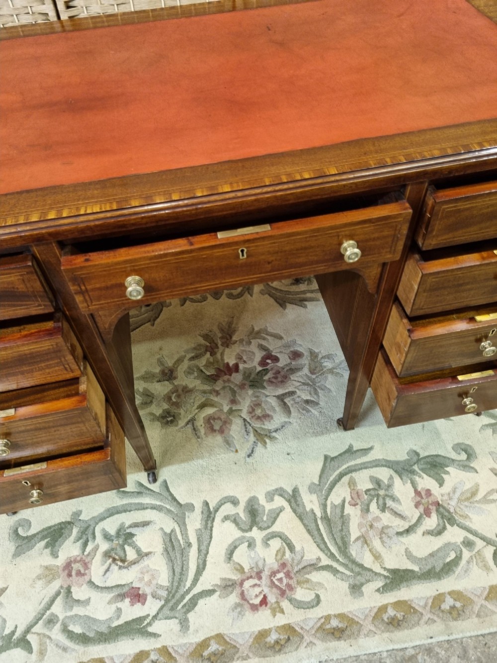 An Edwardian mahogany writing desk featuring nine inlaid drawers, a terracotta leather writing surface, brass knob handles, and tapered legs on castors. View IV