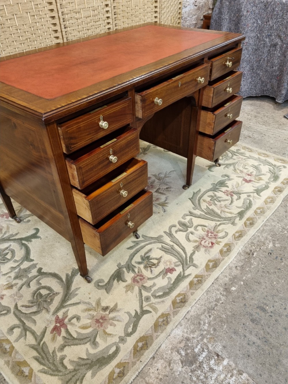 An Edwardian mahogany writing desk featuring nine inlaid drawers, a terracotta leather writing surface, brass knob handles, and tapered legs on castors. View V