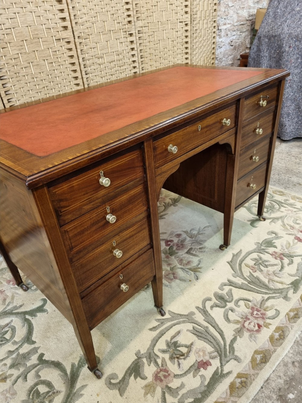 An Edwardian mahogany writing desk featuring nine inlaid drawers, a terracotta leather writing surface, brass knob handles, and tapered legs on castors. View VI