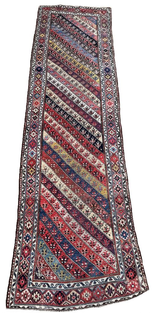 fig 1. A long hand-knotted antique Caucasian wool runner rug featuring diagonal multicoloured geometric bands and intricate floral border motifs.