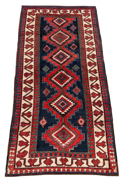 fig 1. An antique Caucasian Kazak wool rug featuring repeating geometric diamond medallions in red, navy blue, green, and ivory framed by stepped borders.