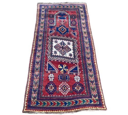 fig 1. A hand-knotted Caucasian prayer rug featuring a red central field with a pointed mihrab niche, geometric medallions, and multi-band patterned borders in navy blue, red, and cream.