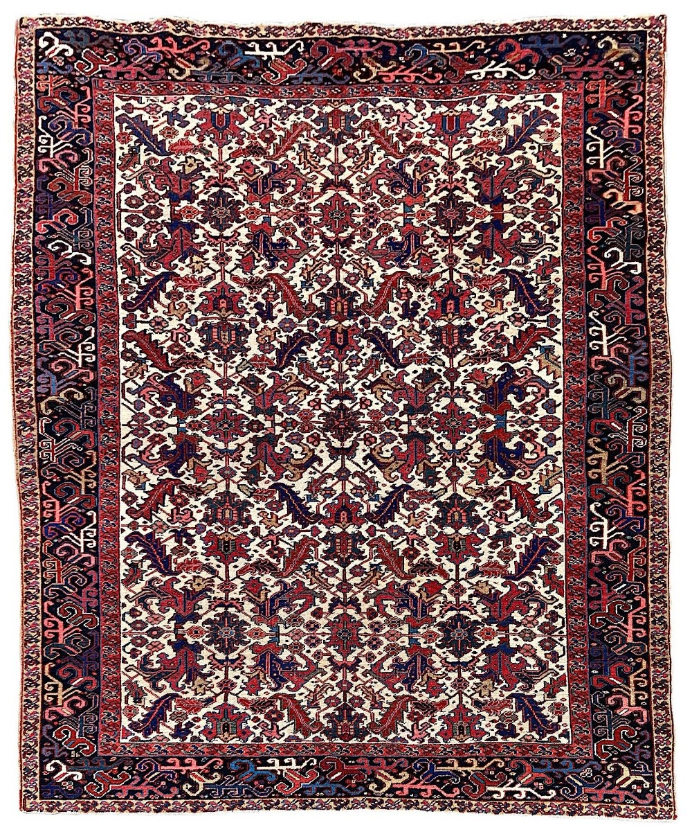 An antique hand-knotted Heriz carpet featuring an ivory central field filled with red and navy geometric floral motifs and framed by a dark blue patterned border.