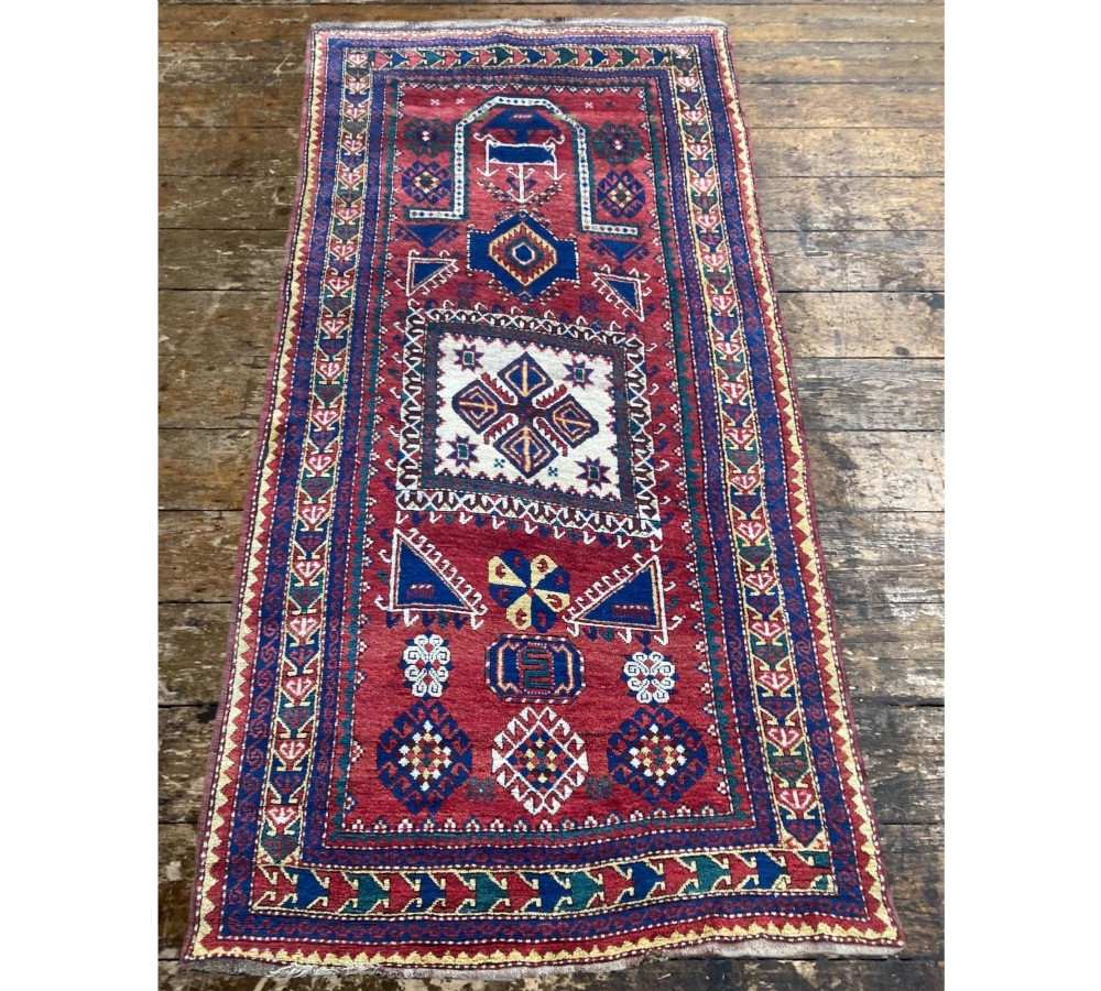 A hand-knotted Caucasian prayer rug featuring a red central field with a pointed mihrab niche, geometric medallions, and multi-band patterned borders in navy blue, red, and cream. View IV